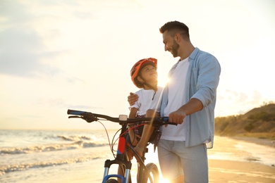 Happy father teaching son to ride bicycle on sandy beach near sea at sunset