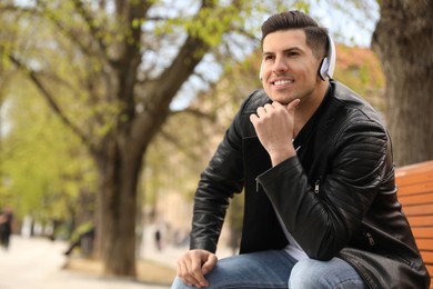 Photo of Handsome man with headphones listening to music while sitting on bench in park, space for text
