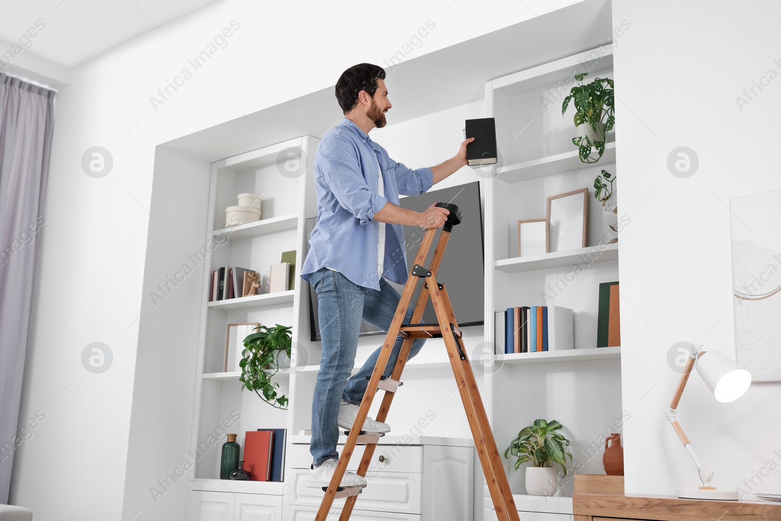 Photo of Man on wooden folding ladder taking books from shelf at home