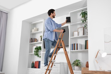 Photo of Man on wooden folding ladder taking books from shelf at home