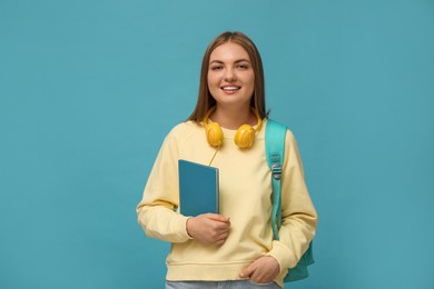 Photo of Teenage student with headphones, backpack and book on light blue background