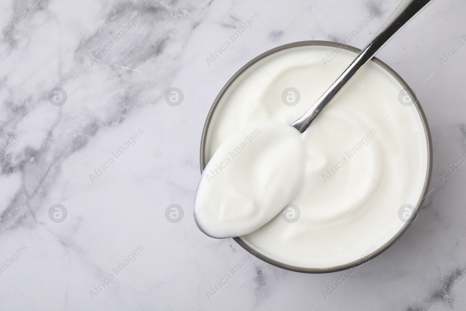 Photo of Delicious natural yogurt in bowl and spoon on white marble table, top view. Space for text