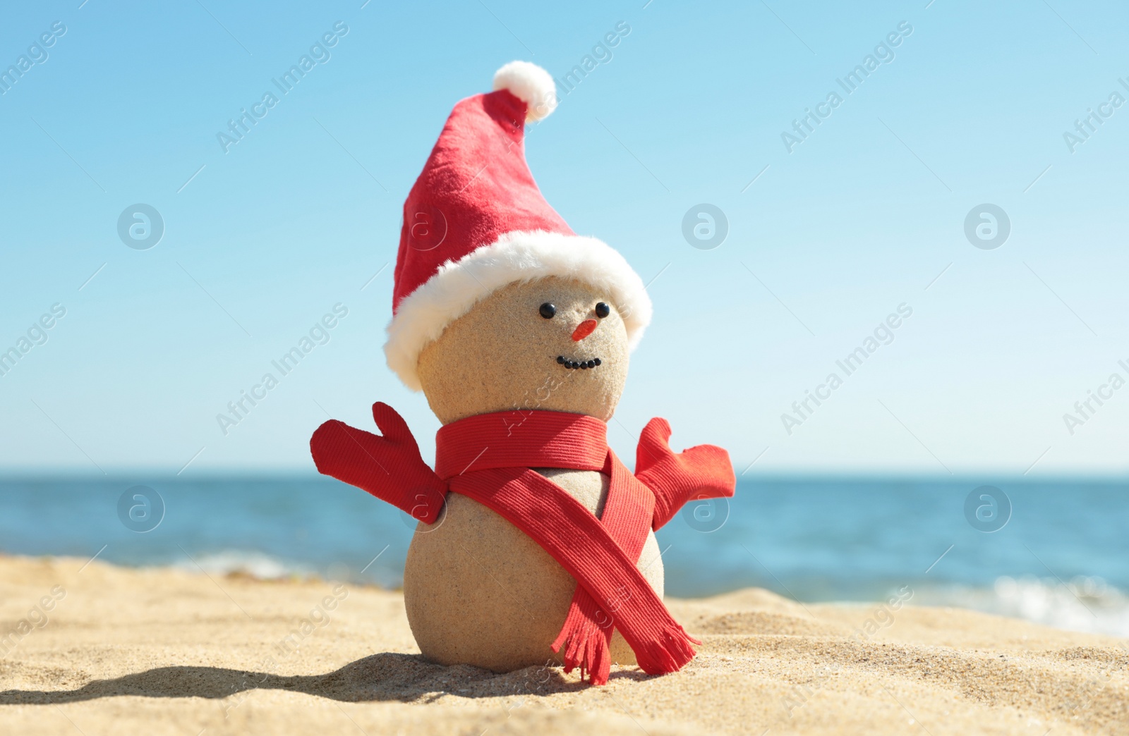 Photo of Snowman made of sand with Santa hat and scarf on beach near sea. Christmas vacation