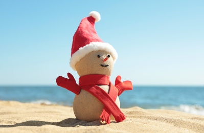 Photo of Snowman made of sand with Santa hat and scarf on beach near sea. Christmas vacation