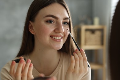 Photo of Smiling woman with freckles applying makeup near mirror indoors