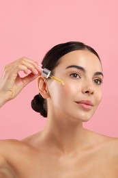 Beautiful young woman applying serum onto her face on pink background