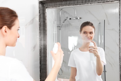 Photo of Young woman applying thermal water on face near mirror in bathroom. Cosmetic product