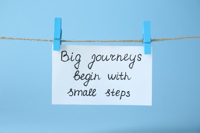 Photo of Card with phrase Big Journeys Begin With Small Steps hanging on rope against light blue background. Motivational quote