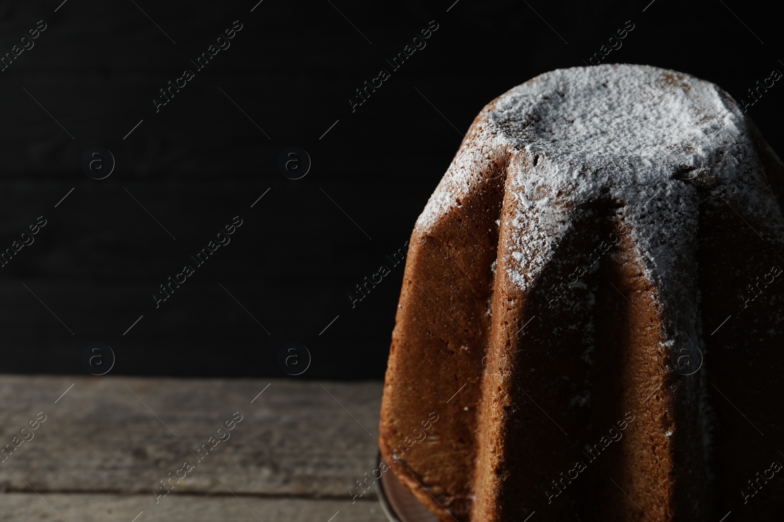 Photo of Delicious Pandoro cake decorated with powdered sugar on wooden table against black background, closeup and space for text. Traditional Italian pastry