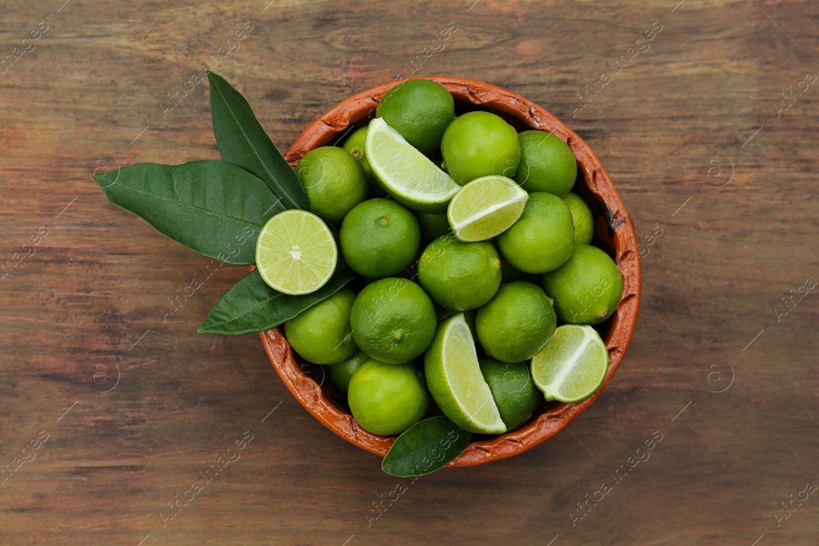 Photo of Whole and cut fresh ripe limes in bowl on wooden table, top view