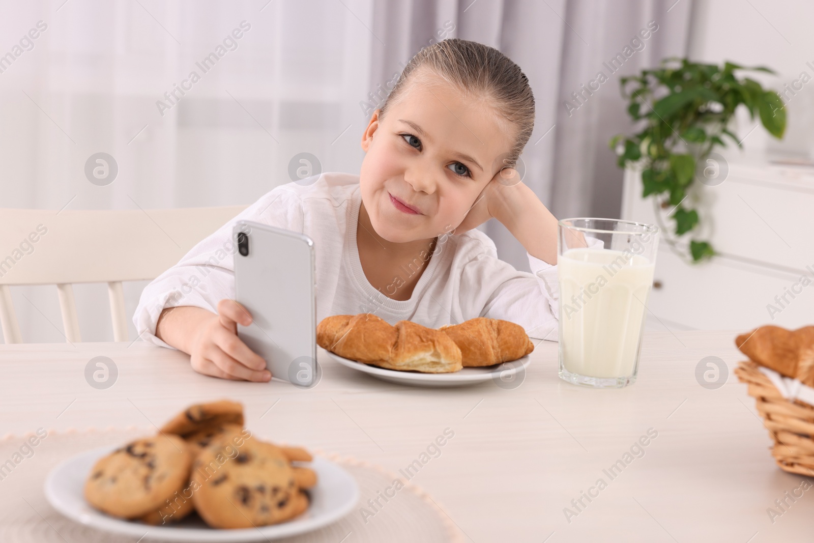 Photo of Little girl using smartphone while having breakfast at table indoors. Internet addiction