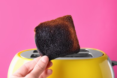 Photo of Woman holding burnt bread near toaster against pink background, closeup