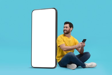 Man with mobile phone sitting near huge device with empty screen on light blue background. Mockup for design