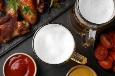 Photo of Mugs with beer, delicious baked chicken wings and sauces on dark grey table, flat lay