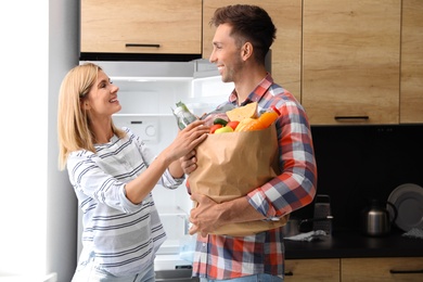 Photo of Happy couple with paper bag full of products near refrigerator in kitchen