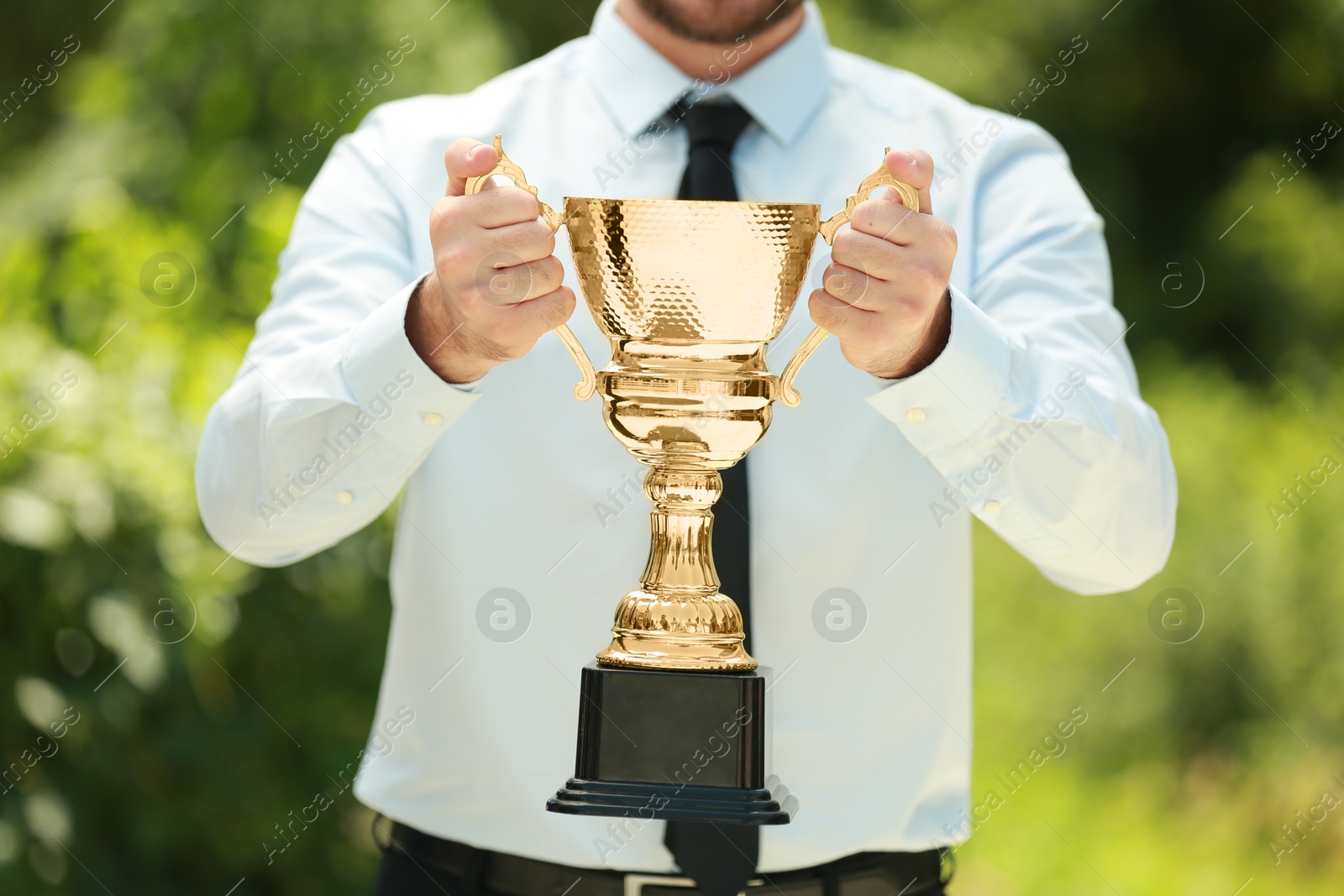 Photo of Young businessman holding gold trophy cup in green park, closeup