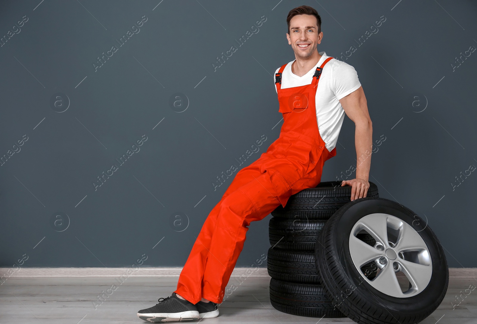 Photo of Young mechanic sitting on stack of car tires near dark wall