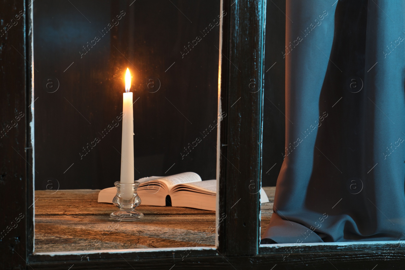 Photo of Burning candle and Bible on wooden table at night, view through window