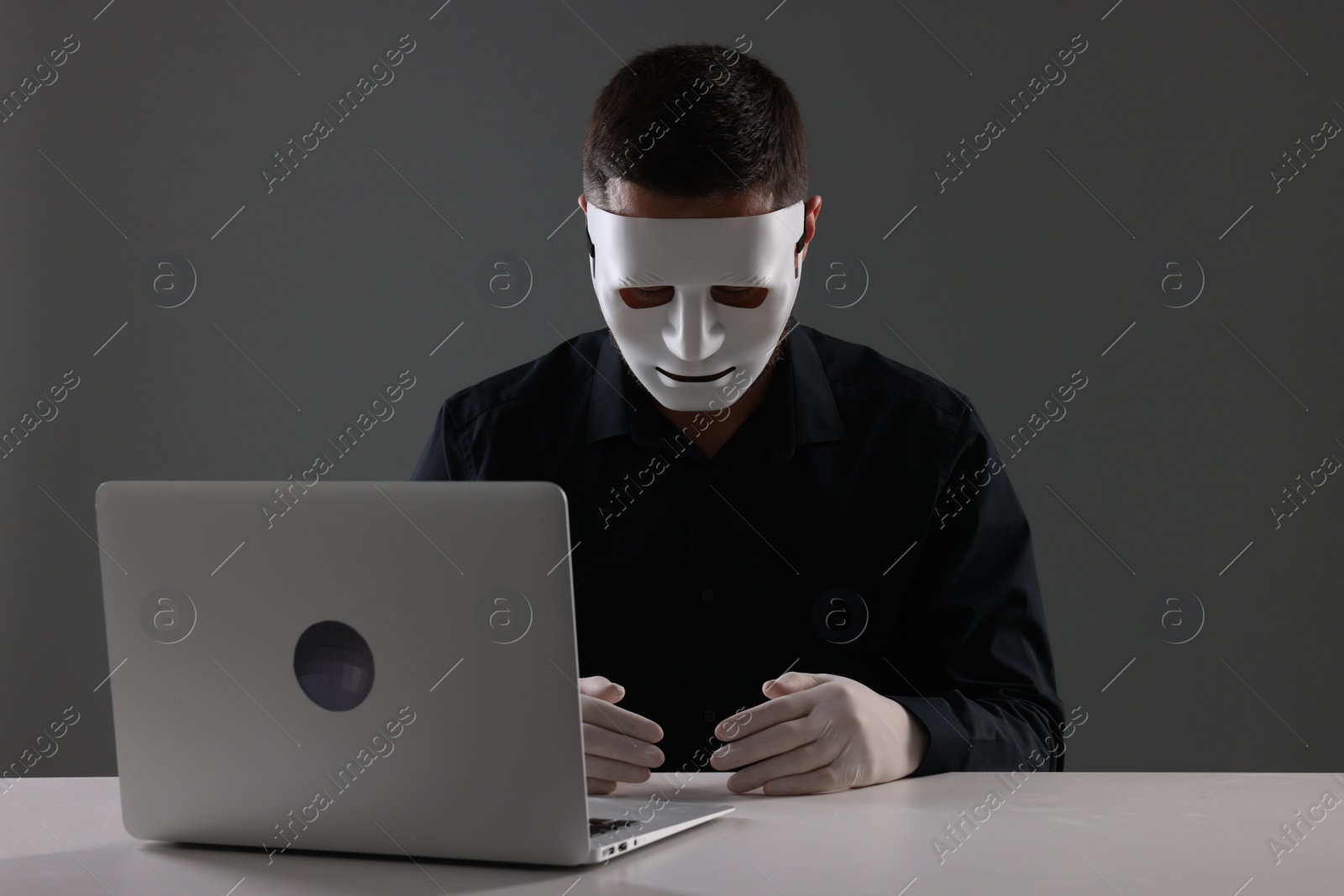 Photo of Man in mask and gloves sitting with laptop at white table against grey background