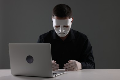 Man in mask and gloves sitting with laptop at white table against grey background