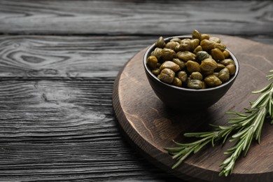 Photo of Tasty capers in bowl and rosemary on black wooden table, space for text