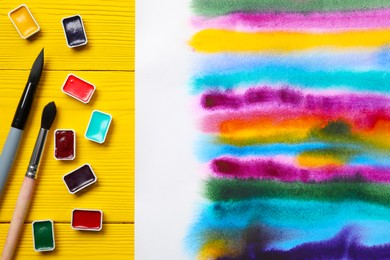 Photo of Bright abstract watercolor painting, colorful paints, and brushes on yellow wooden table, flat lay