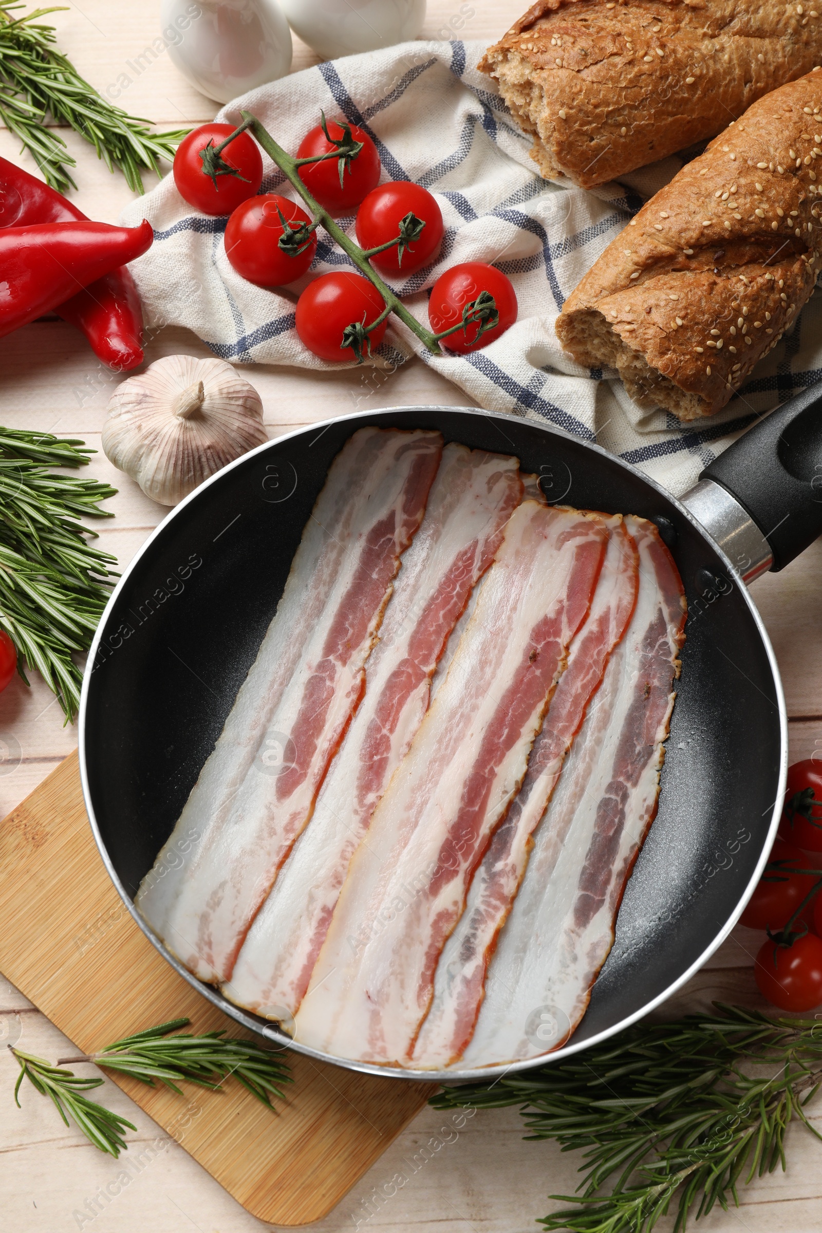 Photo of Slices of raw bacon in frying pan and fresh products on wooden table, flat lay