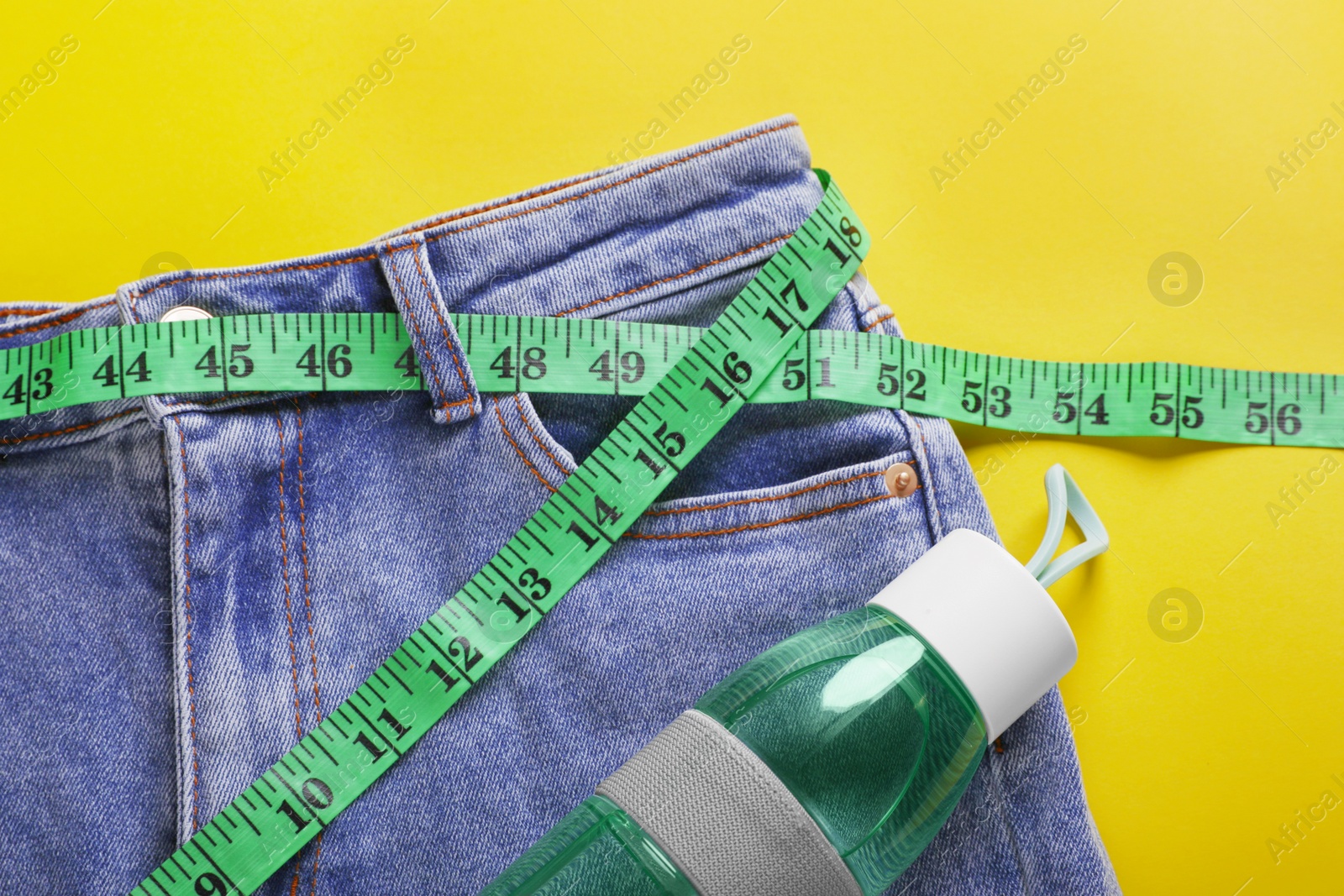 Photo of Jeans with measuring tape and bottle of water on yellow background, flat lay. Weight loss concept