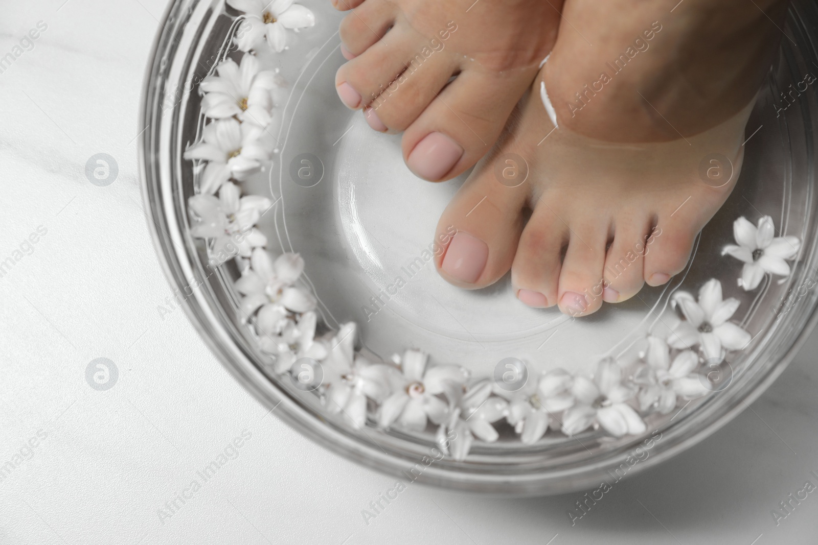 Photo of Woman soaking her feet in bowl with water and flowers on white marble table, top view. Pedicure procedure