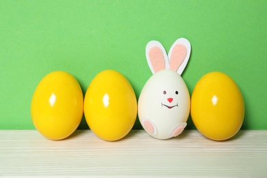 Photo of Bright Easter eggs and white one as cute bunny on wooden table against green background