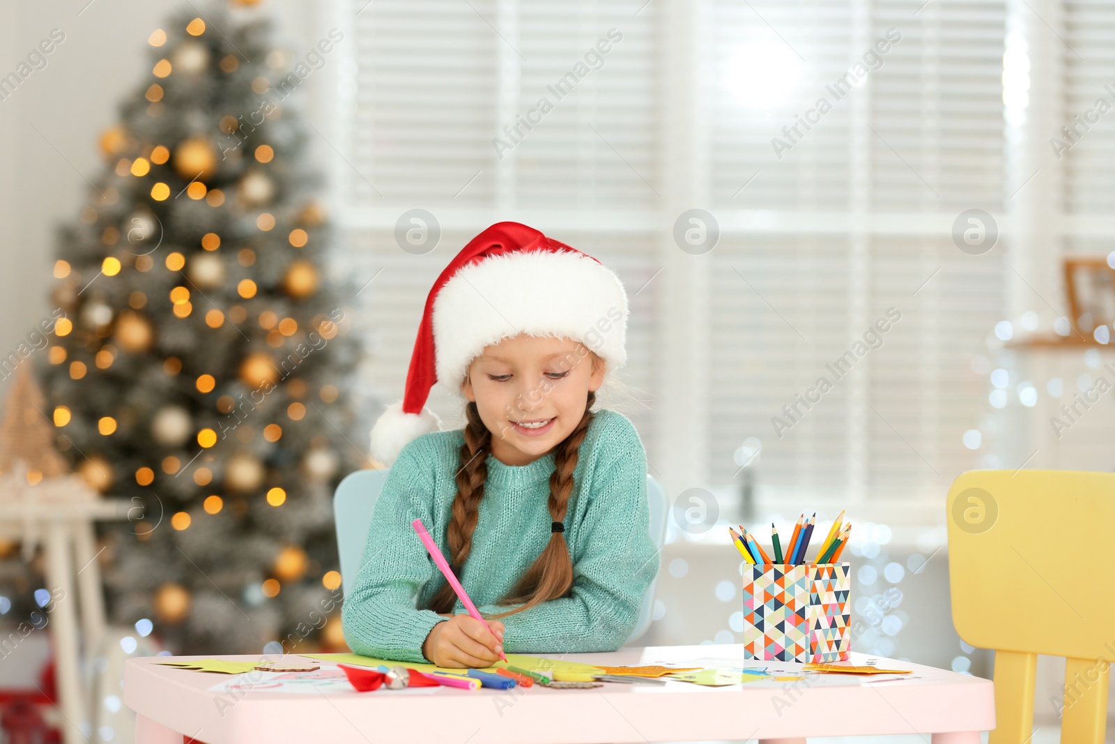 Photo of Little child in Santa hat drawing at table indoors. Christmas season