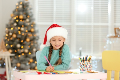 Little child in Santa hat drawing at table indoors. Christmas season