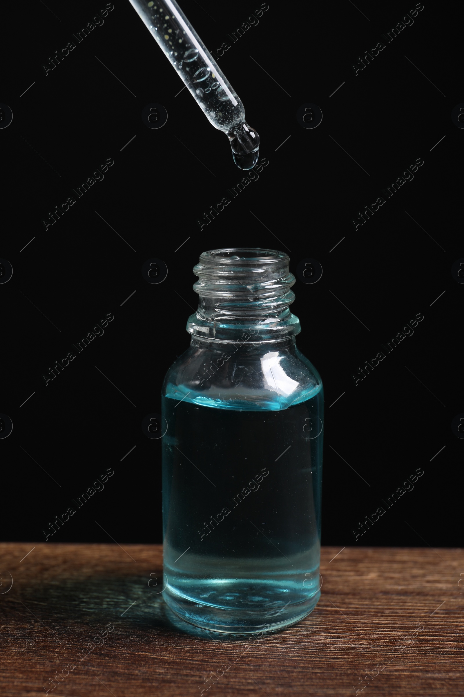 Photo of Dripping cosmetic serum from pipette into bottle at wooden table against black background