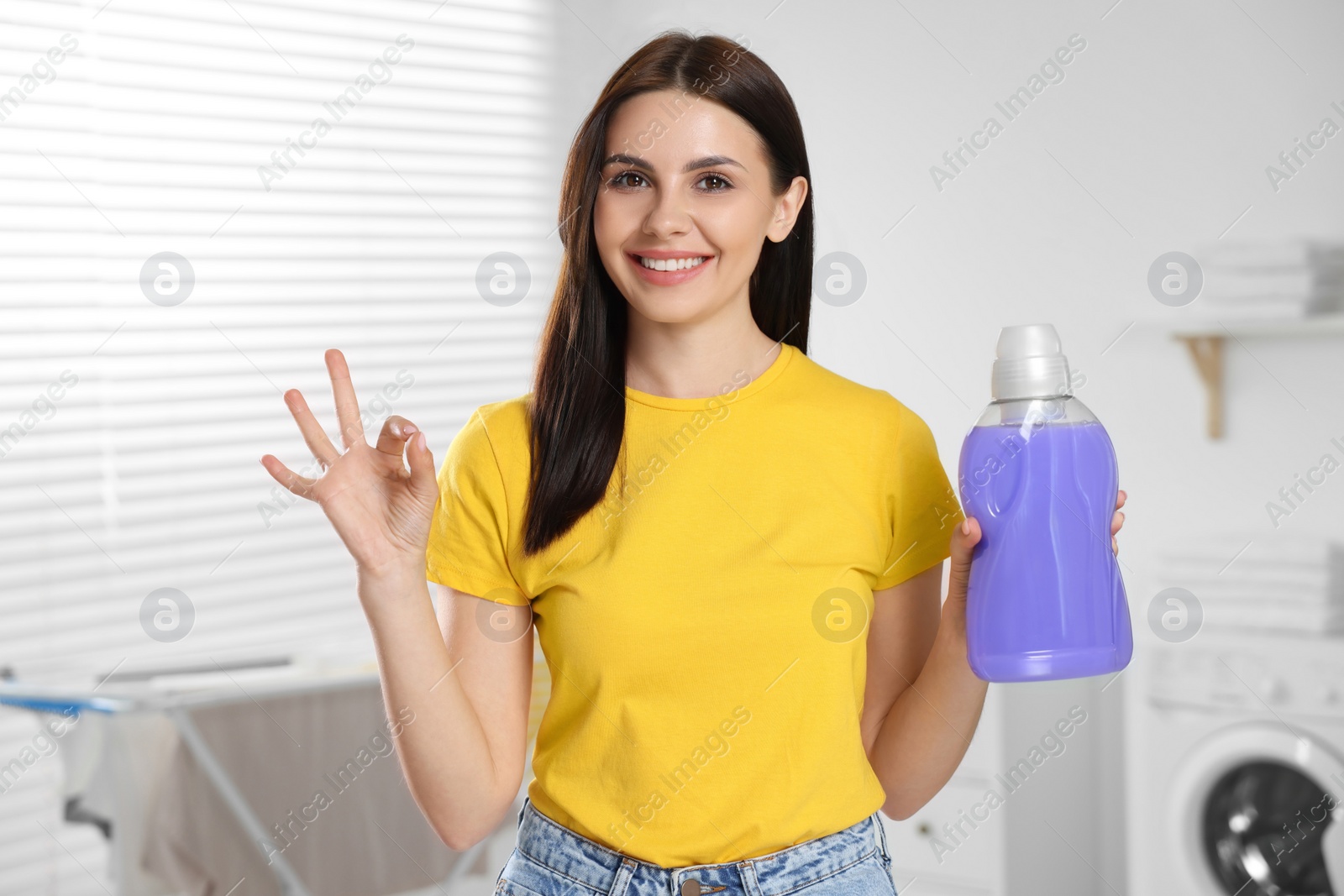 Photo of Woman holding fabric softener and showing OK gesture in bathroom