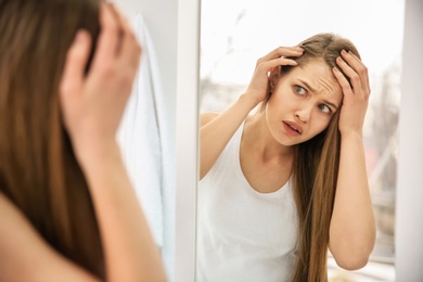 Young woman with hair loss problem looking in mirror indoors