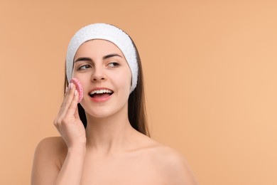 Photo of Young woman with headband washing her face using sponge on beige background, space for text