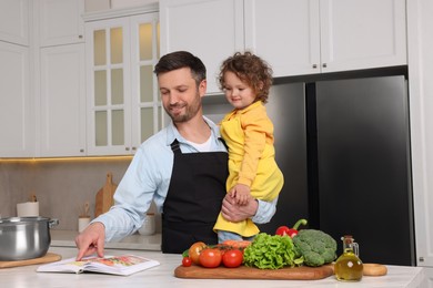 Photo of Cute little girl and her father with recipe book cooking in kitchen