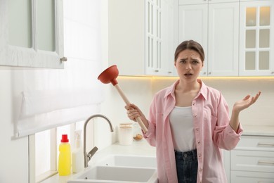 Photo of Upset young woman with plunger near clogged sink in kitchen
