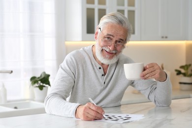 Photo of Senior man with cup of drink solving crossword at table in kitchen