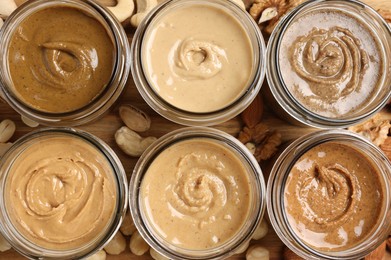 Photo of Many tasty nut butters in jars and nuts on wooden table, flat lay