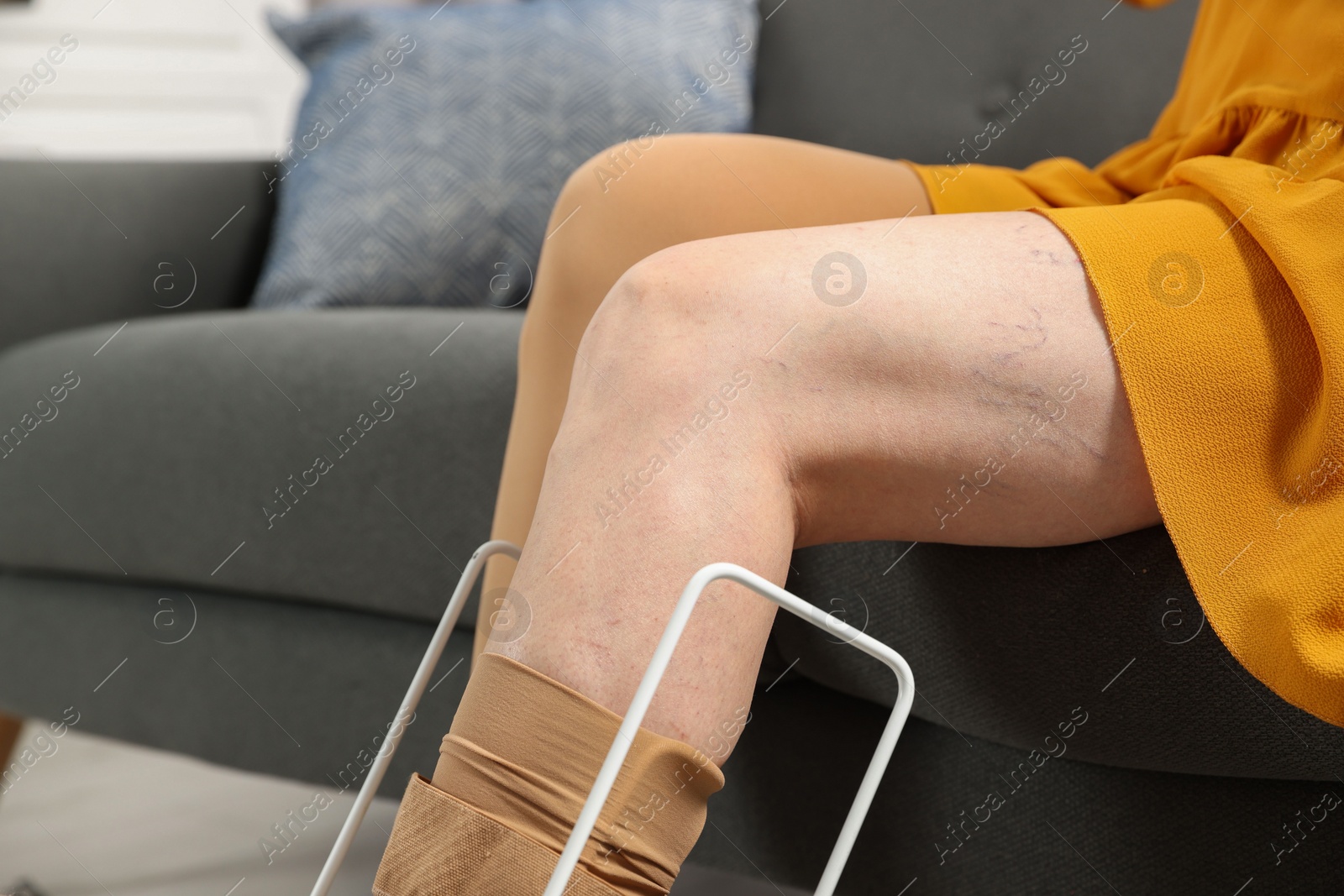 Photo of Woman putting on compression tights with stocking donner in living room, closeup. Prevention of varicose veins