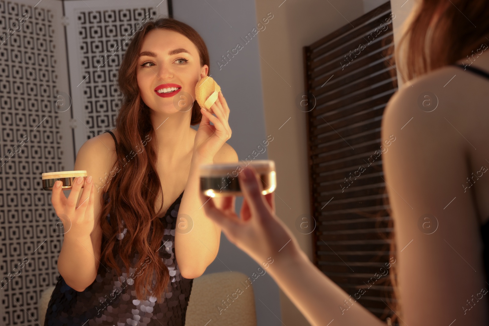 Photo of Beautiful young woman in elegant dress applying powder with puff applicator near mirror indoors