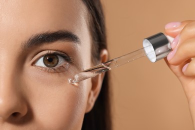 Young woman applying serum onto her face on beige background, closeup