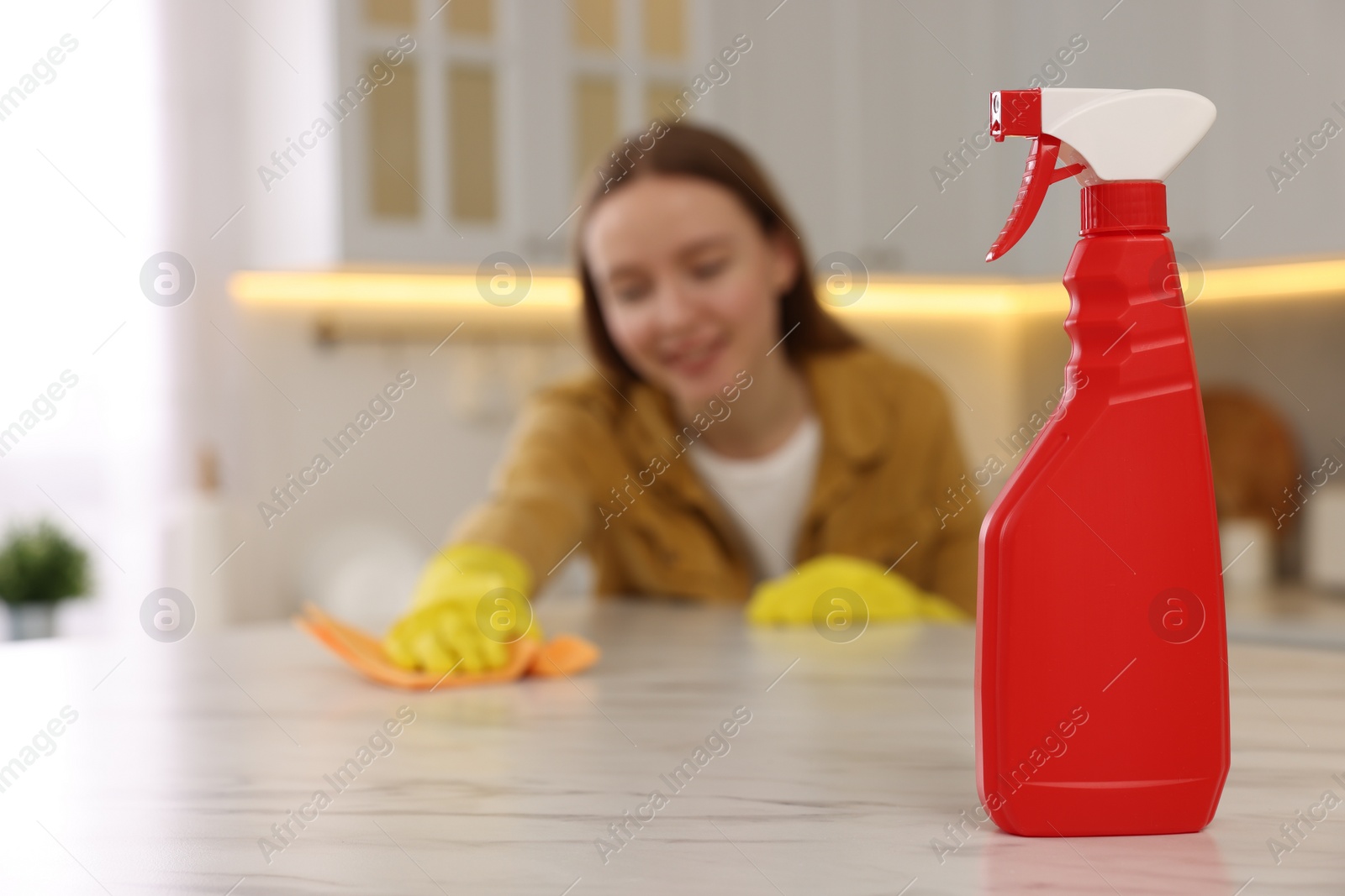 Photo of Woman with microfiber cloth cleaning white marble table in kitchen, focus on spray bottle