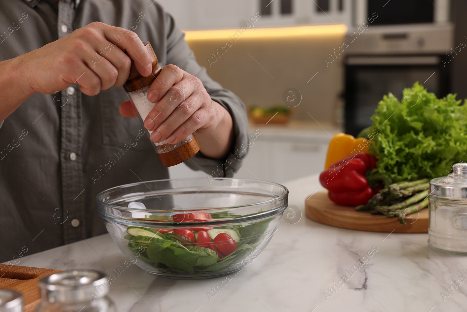 Photo of Cooking process. Man adding salt into bowl of salad at white marble countertop kitchen, closeup