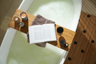 Photo of Wooden bath tray with open book, glass of wine and cosmetic products on tub indoors, top view