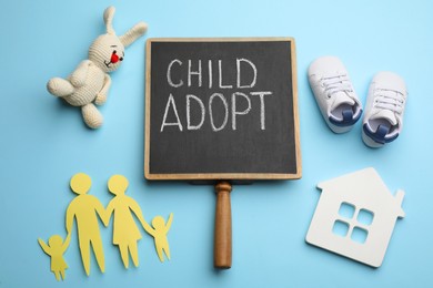Photo of Small chalkboard with phrase CHILD ADOPT, toys, baby shoes and paper family figure on light blue background, flat lay