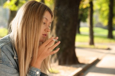 Photo of Young woman smoking cigarette in park on sunny day, space for text