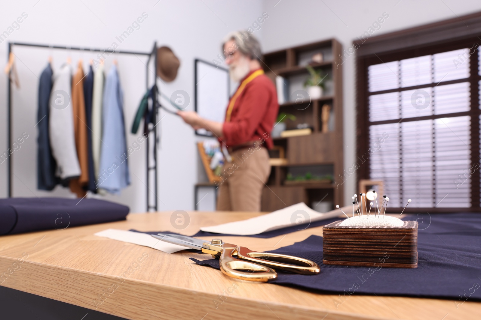 Photo of Professional tailor working in atelier, focus on table with scissors and pin cushion. Space for text