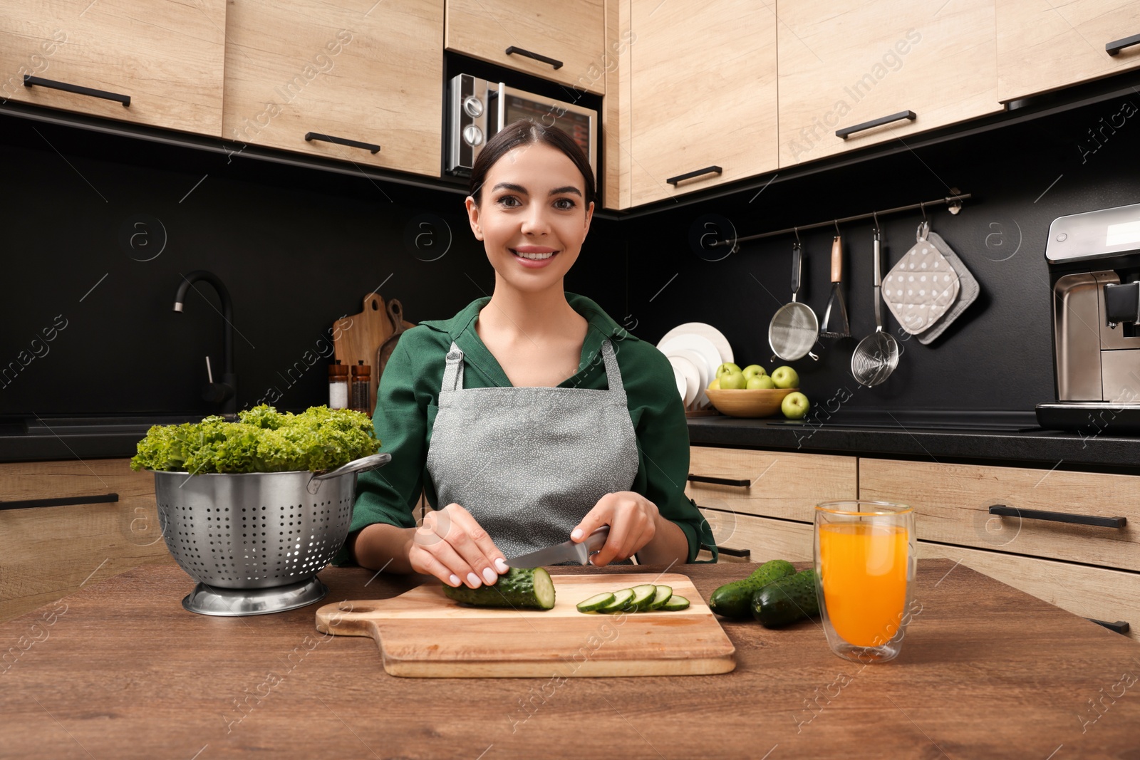 Photo of Woman in apron cutting cucumber at wooden table indoors